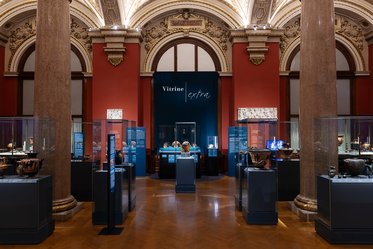 Eine Innenansicht eines Museums mit mehreren Vitrinen, die antike Kunstwerke und Artefakte zeigen. Die Wände sind rot und die Decke ist reich verziert. In der Mitte steht ein großer Schriftzug "Vitrine extra". Das Licht beleuchtet die Objekte in den Glaskästen.