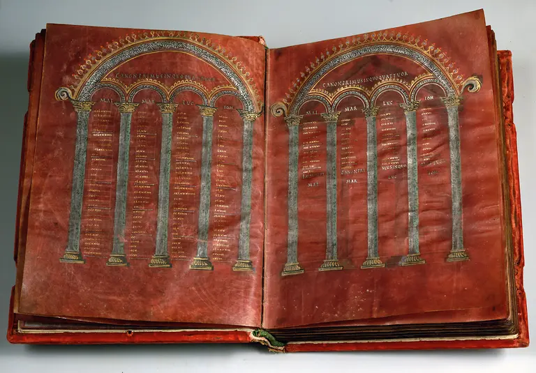 Das Bild zeigt eine geöffnete, alte Buchseite mit aufwendig gestalteten Säulen und einer verzierte Bogen. In den Säulen stehen beschriftete Textzeilen. Der Einband des Buches ist rot und zeigt an den Rändern eine detaillierte Verzierung.