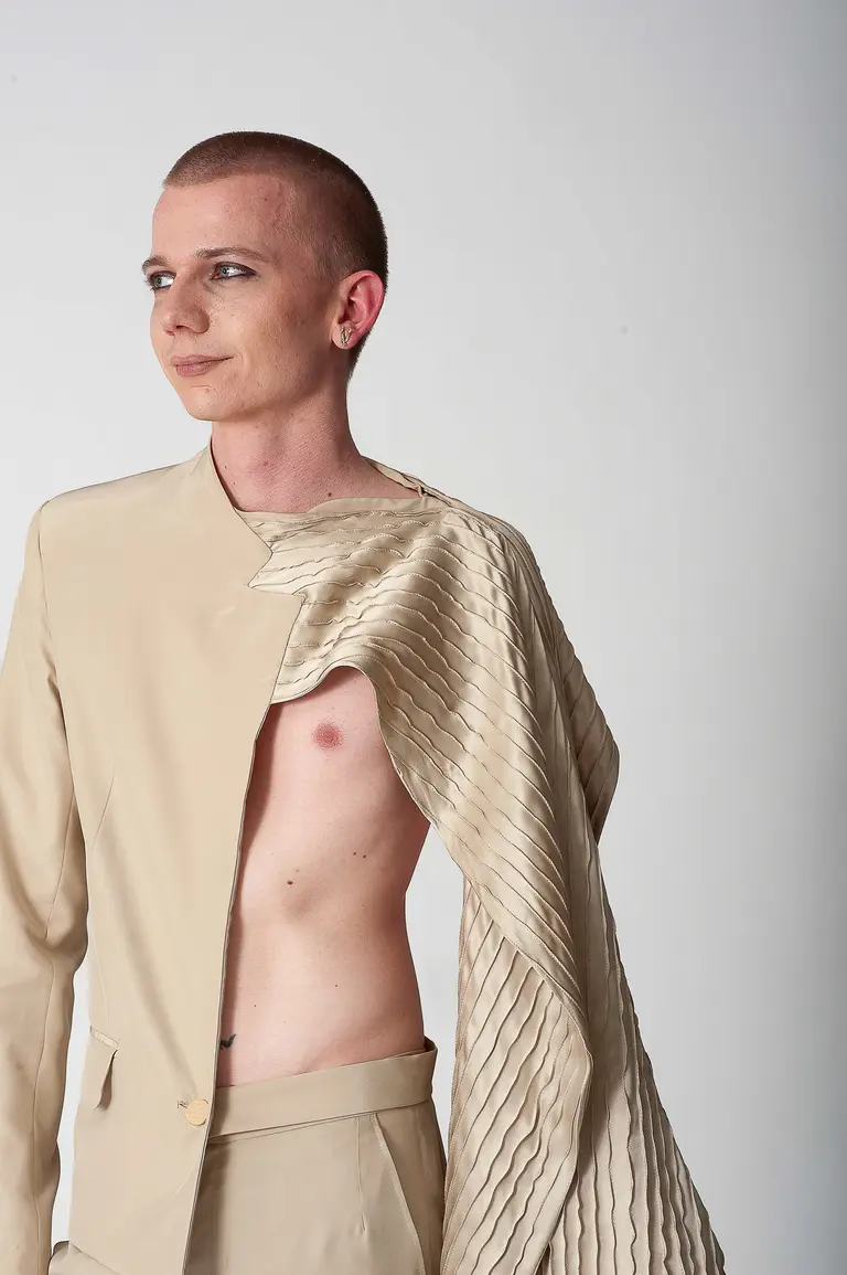 A person with short hair stands confidently, wearing a unique, beige outfit that features an asymmetrical design. One side of the jacket is draped elegantly while the other side is open, revealing part of their torso. The background is plain and bright.