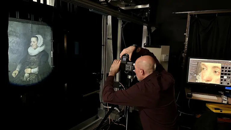 A person is using a camera to photograph a classic portrait displayed on a screen. The portrait features a woman in historical attire with a white collar. The setting appears to be a studio with dim lighting, focused on the art capture process.