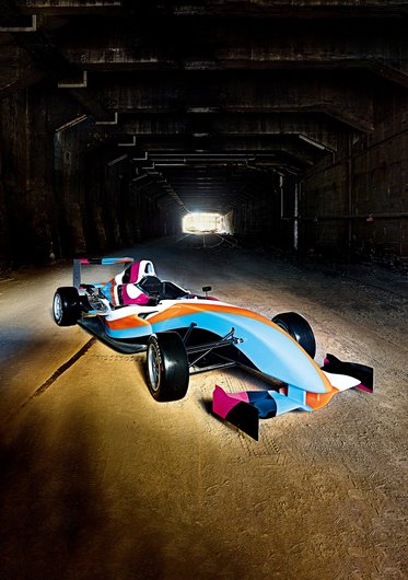 Ein farbenfrohes Formelauto steht auf einer staubigen Piste in einem Tunnel. Der Tunnel ist dunkel, während am Ende ein helles Licht leuchtet. Das Auto hat auffällige Farben in Blau, Pink und Orange und zeigt sportliche Merkmale.
