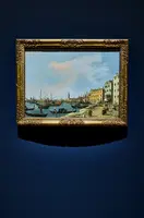 Ein gemaltes Landschaftsbild mit einem Hafen, vielen Schiffen und Gebäuden am Ufer. Der Himmel ist blau mit einigen Wolken. Das Bild ist in einem goldenen Rahmen präsentiert und hängt an einer dunklen Wand.