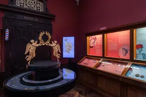 Im Vordergrund steht ein runder Sockel mit einer Skulptur von zwei Engeln, die einen Lorbeerkranz halten. Im Hintergrund sieht man Vitrinen mit historischen Exponaten und Textinformationen, alles in einem rahmenden, gemütlichen Ausstellungsraum mit roten Wänden.