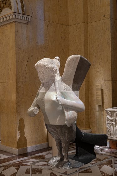 Die Skulptur zeigt eine geflügelte Figur, die in einer dynamischen Pose dargestellt ist. Der obere Teil besteht aus weißem Material, während der untere Teil aus dunklerem Stein gefertigt ist. Die Figur hat ein menschliches Antlitz und hält einen Gegenstand in der Hand.