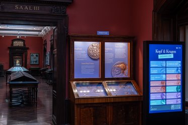 Im Bild ist ein Museumsausstellungsraum mit rot gestrichenen Wänden zu sehen. An der Wand sind Informationsschilder und Vitrinen mit Exponaten, einschließlich Münzen. Auf der rechten Seite befindet sich ein interaktiver Bildschirm mit dem Titel „Kopf & Krugen“.