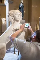 Ein Besucher fotografiert eine antike Skulptur mit mehreren Köpfen in einem Museum. Im Hintergrund sind weitere Kunstwerke zu sehen. Die Szene vermittelt eine Atmosphäre der Wertschätzung für historische Kunst.