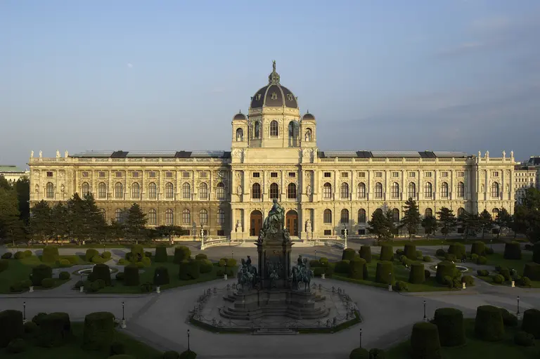 Das Bild zeigt die prächtige Fassade eines historischen Museums mit einer großen Kuppel. Im Vordergrund befindet sich ein schmuckvoller Brunnen und gepflegte Grünflächen mit Sträuchern. Der Himmel ist klar und die Sonne beleuchtet die Architektur.