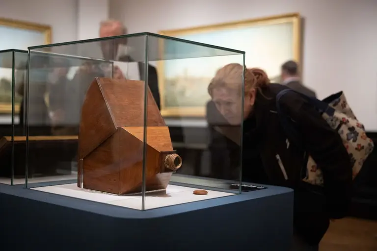 Eine Person beugt sich über ein ausgestellten Holzobjekt in einem Glasdisplay. Das Objekt hat eine geometrische Form und einen runden Ausschnitt. Im Hintergrund sind verschwommene Besucher und Gemälde zu sehen.