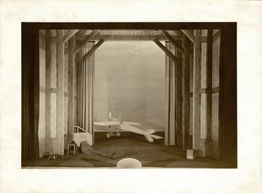 Das Bild zeigt eine Flugzeughalle mit einem weißen Flugzeug im Vordergrund. Im Hintergrund sind große Holzbalken und Vorhänge zu sehen. Links stehen ein Tisch und ein Stuhl. Die Szene vermittelt eine Atmosphäre der Vorbereitung und des Wartens auf den Flug.