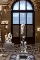 Eine kunstvolle Ausstellung mit verschiedenen Skulpturen aus Marmor und Metall. Im Hintergrund ist ein großes Fenster, das natürliches Licht hereinlässt. Die Skulpturen zeigen menschliche Figuren in unterschiedlichen Posen und Stilen, die in einem eleganten Raum angeordnet sind.
