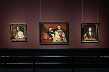 Drei Gemälde hängen in einem Ausstellungsraum. Links ist ein Porträt einer Frau, in der Mitte sind zwei Personen, die eine Schüssel halten, und rechts ein weiteres Porträt einer Frau. Die Wände sind dunkelrot und die Beleuchtung hebt die Kunstwerke hervor.