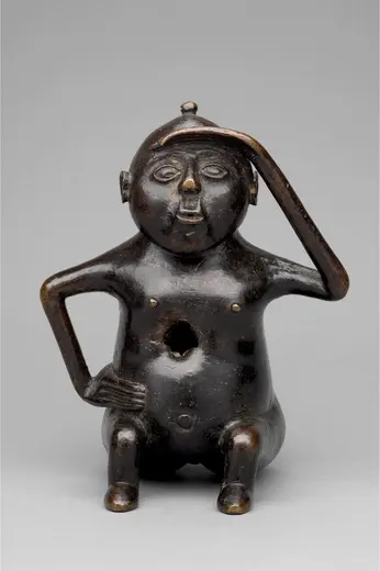 Eine kleine, sitzende Skulptur aus Bronze, die eine menschliche Figur darstellt. Sie hat einen runden Kopf, offene Augen und eine nachdenkliche Pose, mit einer Hand auf der Stirn. Der Körper weist eine Öffnung im Bauchbereich auf, und die Figur sitzt auf einem kleinen Sockel.