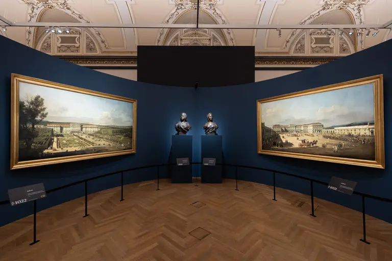 Zwei große Landschaftsgemälde hängen an blauen Wänden, flankiert von zwei Büsten. Der Boden ist aus Holz und die Decke zeigt kunstvolle Details. Die Bilder zeigen verschiedene Landschaften, während die Büsten als Skulpturen in der Mitte des Raumes platziert sind.