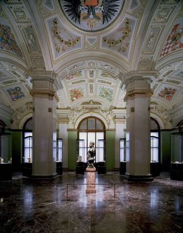 Ein prächtiger Innenraum eines Museums mit hohen Decken und kunstvollen Verzierungen. In der Mitte steht eine Statue auf einem Sockel, umgeben von historischen Objekten in Vitrinen. Die Wände sind in sanften Farben gehalten und Fenster lassen natürliches Licht herein.