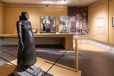 Eine große Statue aus schwarzem Stein steht im Vordergrund. Im Hintergrund sind Vitrinen mit weiteren Artefakten sowie eine Wand mit historischen Bildern und Texten zu sehen. Der Raum ist hell erleuchtet und hat eine moderne Ausstellungsgestaltung.