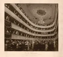 Das Bild zeigt das Innere eines historischen Theaters mit mehreren Rängen. Viele Zuschauer sind auf den Balkonen und im Zuschauerraum zu sehen. Ein Chandelier hängt von der Decke, und eine Person auf der Bühne spricht zu dem Publikum. Die Atmosphäre ist festlich und lebendig.