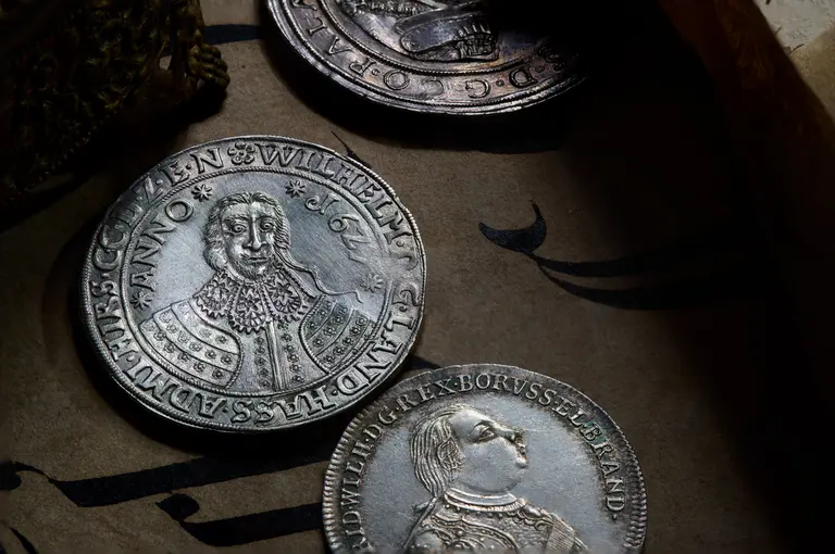 A close-up of several ancient silver coins laid on a textured surface. One coin features a portrait of a historical figure, identified as 