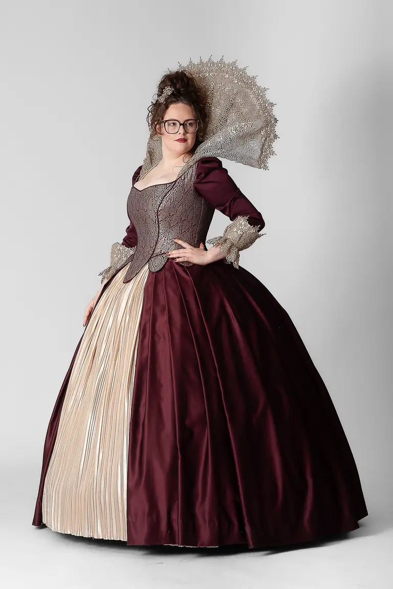 A woman wearing a historical costume stands against a neutral backdrop. The dress features a deep burgundy bodice with intricate detailing and a full, pleated cream skirt. She accessorizes with a decorative silver headdress and glasses, creating a striking, elegant appearance.