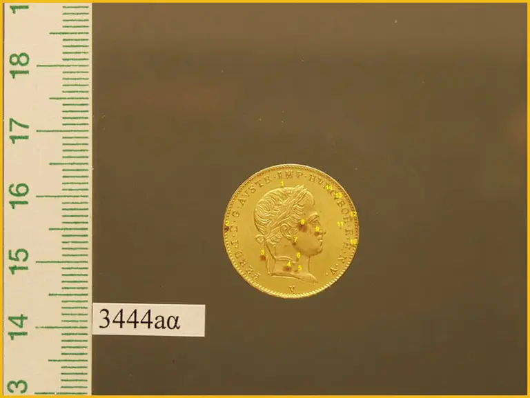 Ein goldener Münze mit einem Profilbild einer Person. Umrandung mit einem Schriftzug und Verzierungen. Links neben der Münze befindet sich ein Maßstab mit Zahlen.