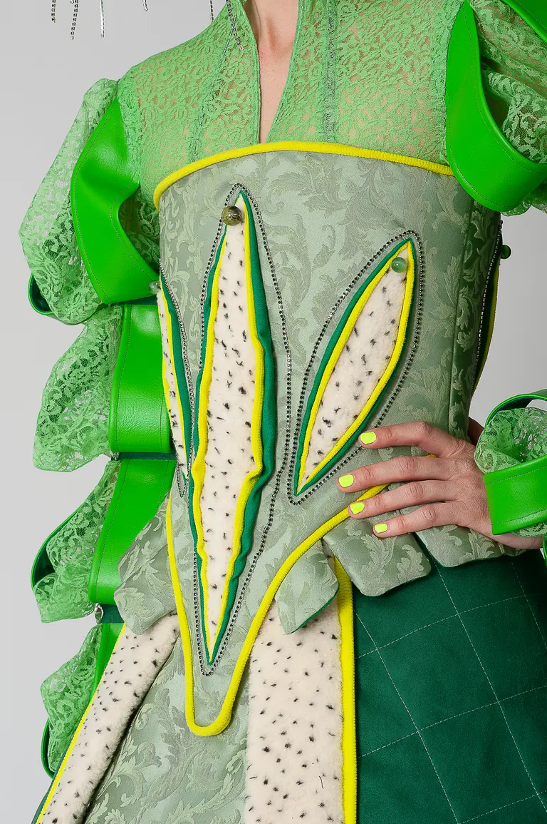 A close-up of a unique fashion design featuring a structured dress with green and yellow details. The bodice has large leaf-like patterns, with ruffled sleeves and intricate stitching. The model's hand, adorned with bright green nail polish, rests on the waist of the garment.