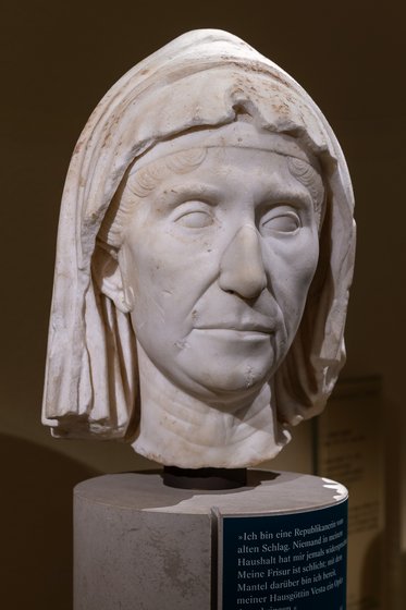 Eine antike Skulptur eines Frauenkopfes aus Marmor. Die Figur hat eine angemessene Frisur und trägt ein Kopftuch. Der Kopf ist auf einem runden Sockel montiert. Im Hintergrund steht eine Informationstafel.