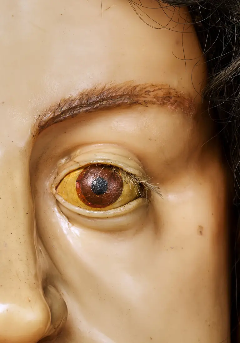 Nahaufnahme eines menschlichen Auges, das aus einer realistischen Wachsfigur stammt. Das Auge hat eine gelb-braune Iris mit einem schwarzen Mittelpunkt. Die Wimpern sind sichtbar, und die Haut um das Auge zeigt leichte Verfärbungen. Der Fokus liegt auf der Detailtreue des Auges.