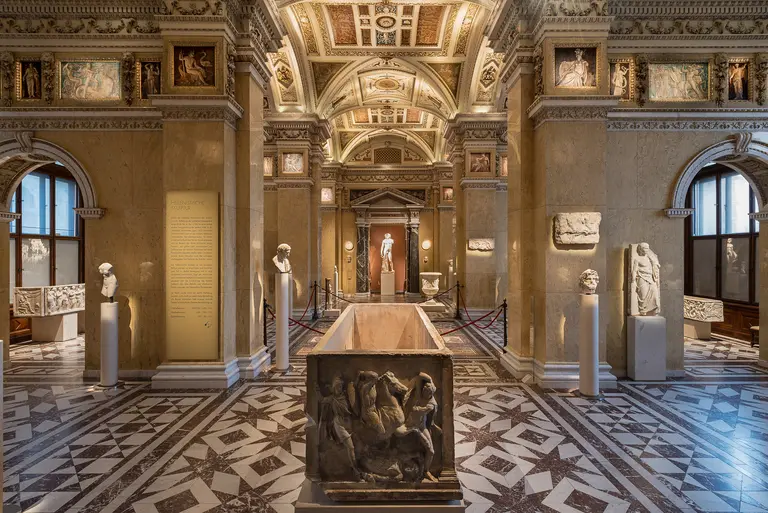 Das Bild zeigt einen eleganten Museumsraum mit kunstvollem Fußboden und hohen Decken. In der Mitte steht ein langer Sarkophag, umgeben von klassischen Statuen und Wandtafeln. Das Licht strömt durch große Fenster, die den Raum erhellen und eine ruhige Atmosphäre schaffen.