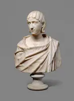 Eine antike Marmorbüste eines jungen Mannes mit lockigem Haar, der ein Tunika ähnliches Gewand trägt. Die Statue ist auf einem runden Sockel platziert und zeigt feine Details im Gesicht und Stoff. Der Hintergrund ist grau.