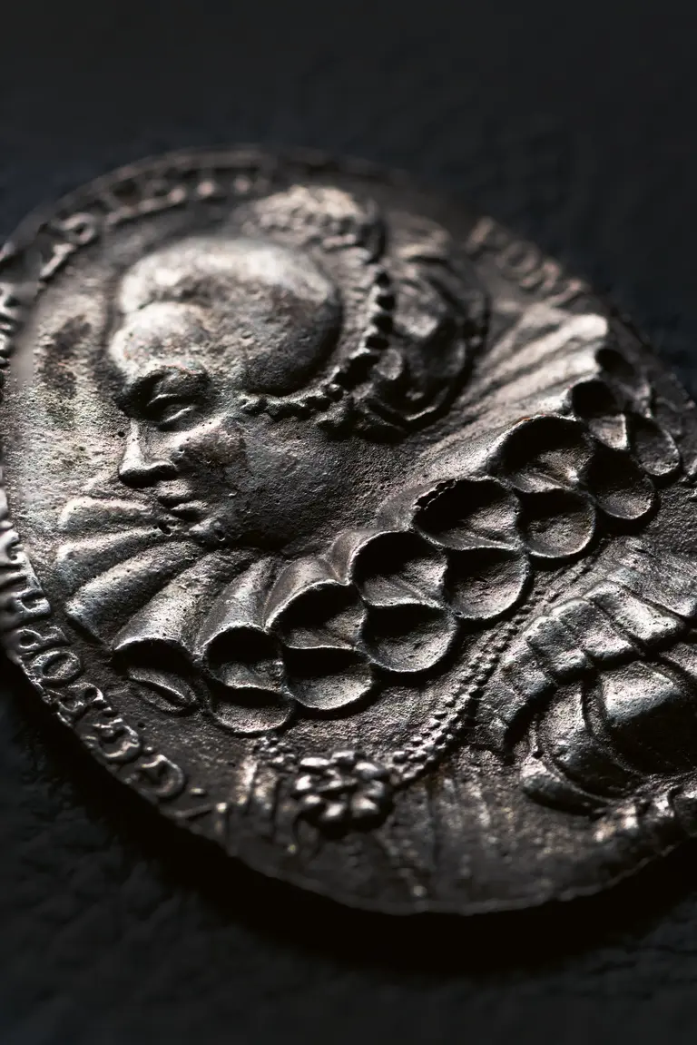 A close-up of an ornate silver medal features a relief of a historical figure's profile, adorned with intricate details such as a ruffled collar. The background is dark, emphasizing the medal's texture and craftsmanship.