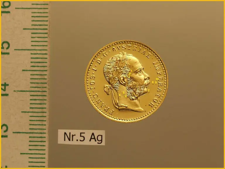 Ein goldener Münze mit einem Portrait eines Herrschers, umgeben von lateinischer Schrift. Die Münze ist auf einem einfarbigen Hintergrund platziert. Links ist ein Maßstab sichtbar. Unter der Münze befindet sich ein kleines Etikett mit der Aufschrift „Nr. 5 Ag“.