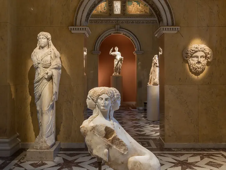 Eine Galerie mit antiken Skulpturen. Links steht eine Statue einer weiblichen Figur, während in der Mitte eine kunstvoll gestaltete Statue eines sitzenden Menschen zu sehen ist. Die Wände sind mit Ornamenten und Malereien dekoriert, die historische Szenen zeigen.