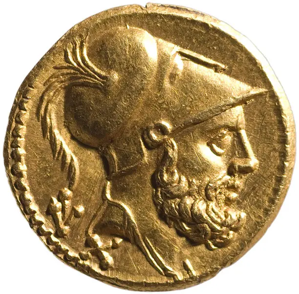 Eine goldene Münze zeigt das Profil eines bärtigen Mannes mit einem Helm, der einen Schopf hat. Die Münze könnte aus der Antike stammen und vermittelt ein Gefühl von Geschichte und kulturellem Erbe.