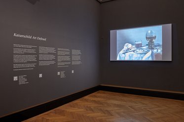 Ein Ausstellungsraum mit einer grauen Wand, auf der Informationen zur "Kaiserschäld Art" zu sehen sind. Auf der gegenüberliegenden Wand befindet sich ein Projektorbild, das ein Stillleben zeigt, mit einer Tischdecke, einem Glas Wasser und verschiedenen Objekten.