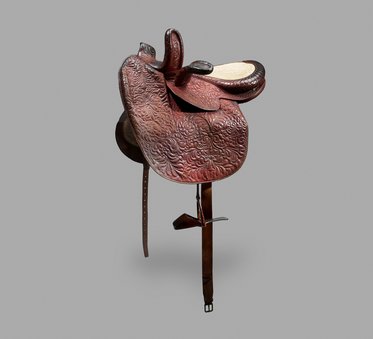 Ein klassischer Westernsattel mit kunstvollen Verzierungen. Der Sattel hat eine dunkle, rissige Lederoberfläche und eine helle Sitzfläche. Er steht aufrecht und zeigt die typischen Merkmale eines Westernsattels, einschließlich der Fender und des Sattelbaums.