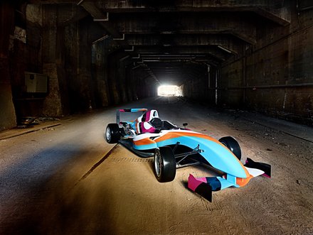 Ein bunter Rennwagen steht in einem dunklen Tunnel. Das Licht am Tunnelende lässt die silberne und blaue Lackierung des Fahrzeugs leuchten. Die Umgebung ist rau und verlassen, wodurch der Fokus auf dem Wagen verstärkt wird.