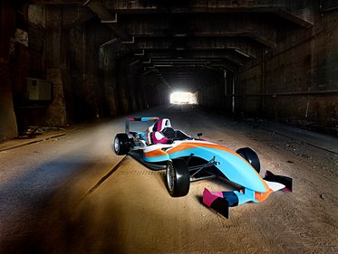 Ein bunter Rennwagen steht in einem dunklen Tunnel. Das Licht am Tunnelende lässt die silberne und blaue Lackierung des Fahrzeugs leuchten. Die Umgebung ist rau und verlassen, wodurch der Fokus auf dem Wagen verstärkt wird.
