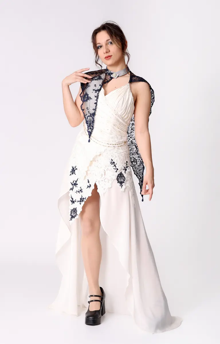 A woman stands elegantly in a stylish white dress with an asymmetrical hemline and lace detailing. She accessorizes with a black lace scarf around her neck and has a confident pose, wearing chunky black heels. The background is plain white, emphasizing her outfit.