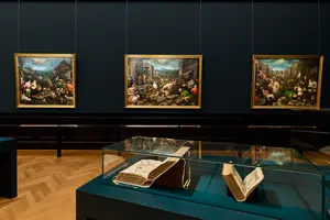 In einem Ausstellungsraum hängen drei große, farbenfrohe Gemälde an der Wand, die lebhafte Marktszenen darstellen. Vor den Bildern befindet sich ein Glaskasten mit einem geöffneten Buch, das Illustrationen zeigt. Der Raum hat einen dunklen Hintergrund und einen hellen Holzboden.