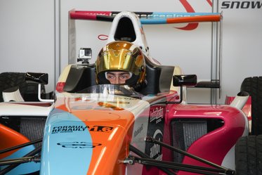 Ein Rennfahrer sitzt in einem offenen Formelwagen. Er trägt einen goldenen Helm und schaut konzentriert nach vorne. Der Wagen hat eine auffällige Lackierung in Blau, Pink und Orange. Im Hintergrund sind Reifenschalen und Teile des Garagenbereichs sichtbar.