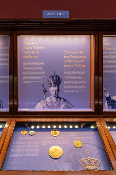 Eine Vitrine mit Informationen zur Krone des Heiligen Römischen Reiches und der Krone von Kaiser Rudolf II. Neben Texten sind goldene Medaillen und Münzen ausgestellt. Oben befindet sich ein Schild mit der Aufschrift „PRESTIGE“.