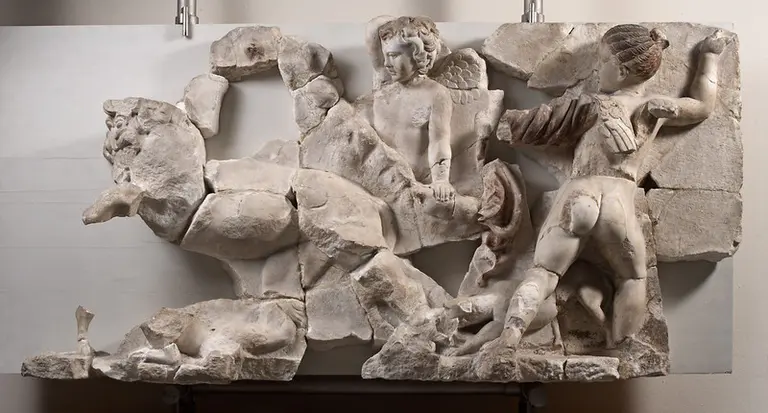 Die Darstellung zeigt ein fragmentiertes Relief, das einen Kämpfer und einen Engel darstellt. Der Kämpfer ist mit einem Schild und einer Rüstung versehen, während der Engel hinter ihm steht und den steigenden Löwen beobachtet. Die Szenen vermitteln einen Kampf zwischen Mensch und Tier.