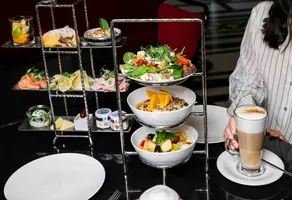Ein mehrstöckiger Ständer mit verschiedenen Speisen, darunter Salat, Obst, Joghurt und Gebäck. Eine Tasse Kaffee steht auf dem Tisch neben einem Teller mit frischen Zutaten. Der Hintergrund ist schattig und elegant.