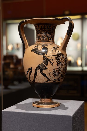 Eine antike griechische Vase steht auf einem Podest. Sie ist aus Ton mit schwarzer Bemalung, die einen reitenden Krieger und florale Muster zeigt. Die Vase hat zwei henkelförmige Griffe und einen runden Fuß. Der Hintergrund ist unscharf, mit weiteren Ausstellungsstücken sichtbar.