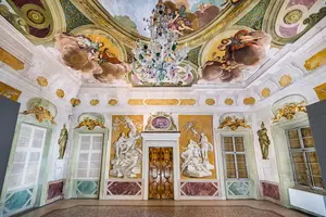 Das Bild zeigt einen kunstvoll gestalteten Raum mit einer prächtigen Deckenmalerei, einem aufwendigen Kronleuchter und symmetrisch angeordneten Wänden, die mit Statuen und dekorativen Elementen verziert sind. Die Farben sind lebhaft und verleihen dem Raum eine elegante Atmosphäre.