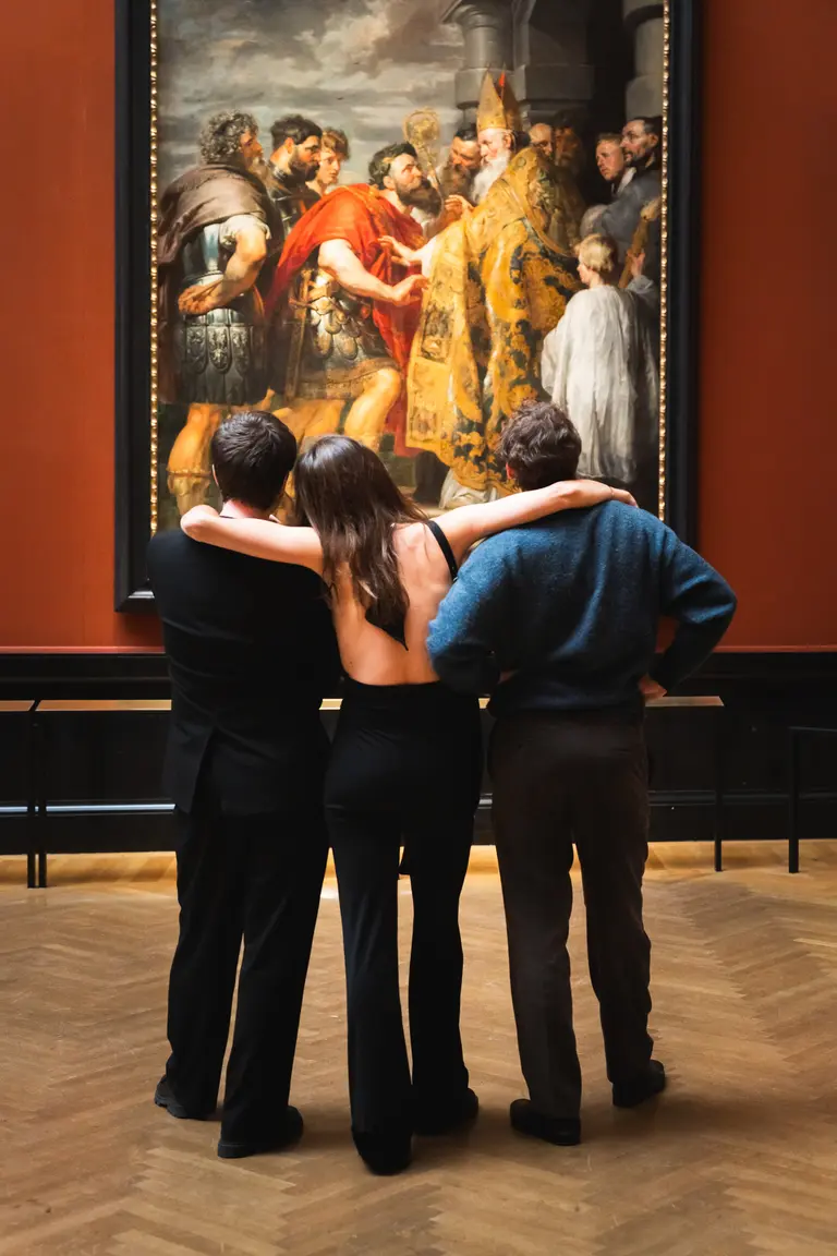 Drei Personen stehen mit dem Rücken zur Kamera und betrachten ein großes Gemälde an der Wand. Die Umgebung ist hell erleuchtet und der Raum wirkt elegant. Die Personen scheinen in das Kunstwerk vertieft zu sein.