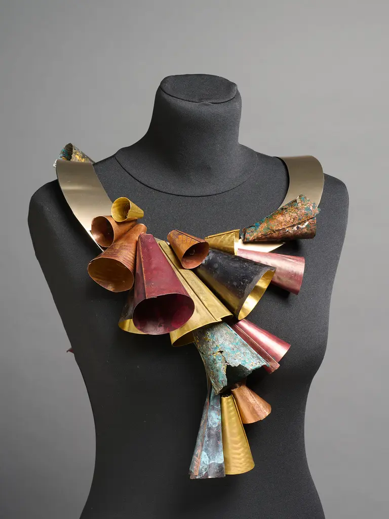 A vibrant, artistic necklace displayed on a black mannequin. The necklace features a variety of metal cones in different colors, including red, gold, and green, arranged in a dynamic, overlapping pattern. The contrasting textures and shapes create a bold statement piece.