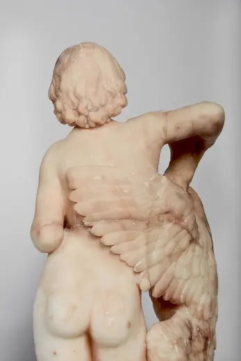 Die Skulptur zeigt eine nackte Figur von hinten, die einen langen, gefiederten Flügel hält. Der Körper ist aus hellem Marmor gearbeitet, mit detailreicher Darstellung der Haare und des Oberkörpers. Der Fokus liegt auf der harmonischen Verbindung zwischen Mensch und Mythos.