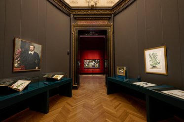 Ein Blick in eine Kunstgalerie mit braunem Holzboden. In den Vitrinen sind Bücher und Illustrationen ausgestellt. An den Wänden hängen ein Porträt eines Mannes und eine Pflanzenzeichnung. Im Hintergrund ist ein weiteres Kunstwerk auf rotem Grund zu sehen.