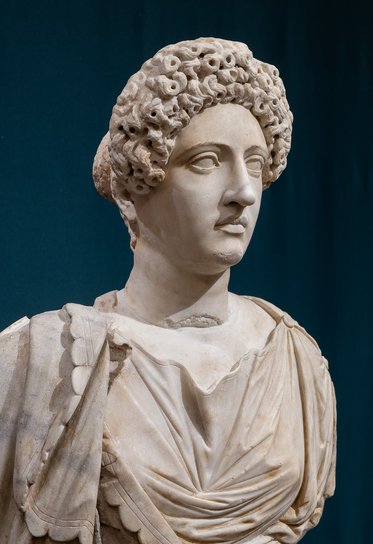 Eine antike Skulptur einer Frau mit lockigem Haar und einer drapierten Tunika. Ihr Blick ist nach vorne gerichtet, und sie hat eine feine, detailreiche Gesichtszüge. Der Hintergrund ist dunkelblau, was die Figur hervorhebt.
