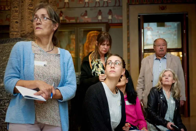Eine Gruppe von fünf Personen in einem Museum. Eine Frau mit einem Notizblock steht vorne, während eine junge Frau mit Brille nachdenklich schaut. Im Hintergrund sind weitere Besucher zu sehen, die verschiedene Stile tragen, vor einer Wand mit antiken Kunstwerken.